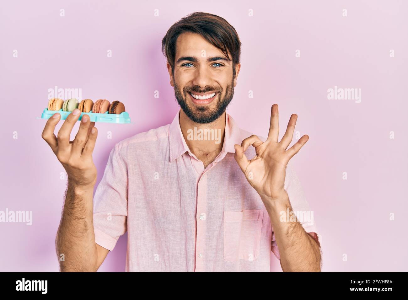 Jeune homme hispanique tenant de délicieux macrons pâtisseries faisant signe ok avec les doigts, souriant sympathique gesturant excellent symbole Banque D'Images