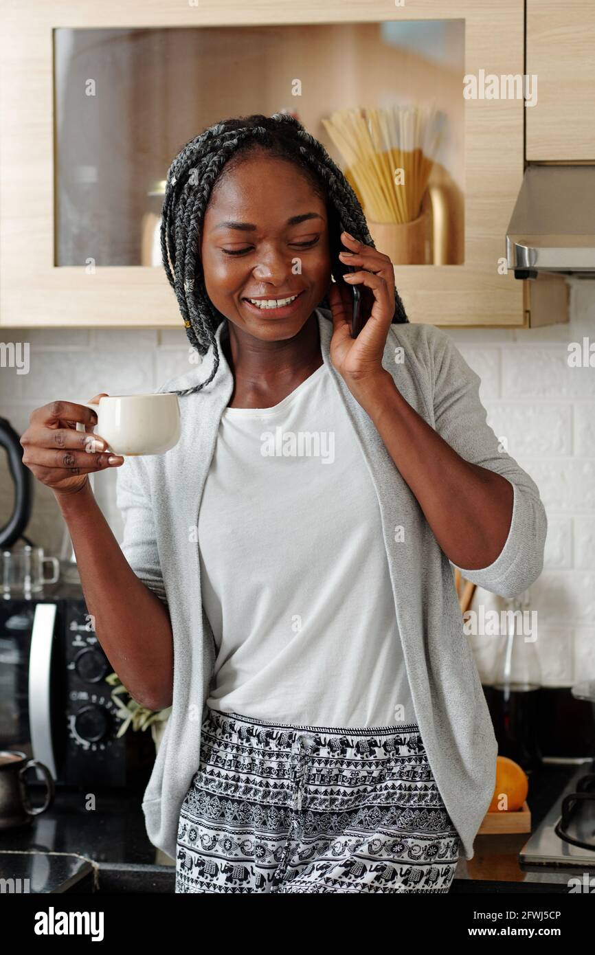 Jolie jeune femme en salon debout dans la cuisine, appelant au téléphone et buvant une tasse de café du matin Banque D'Images