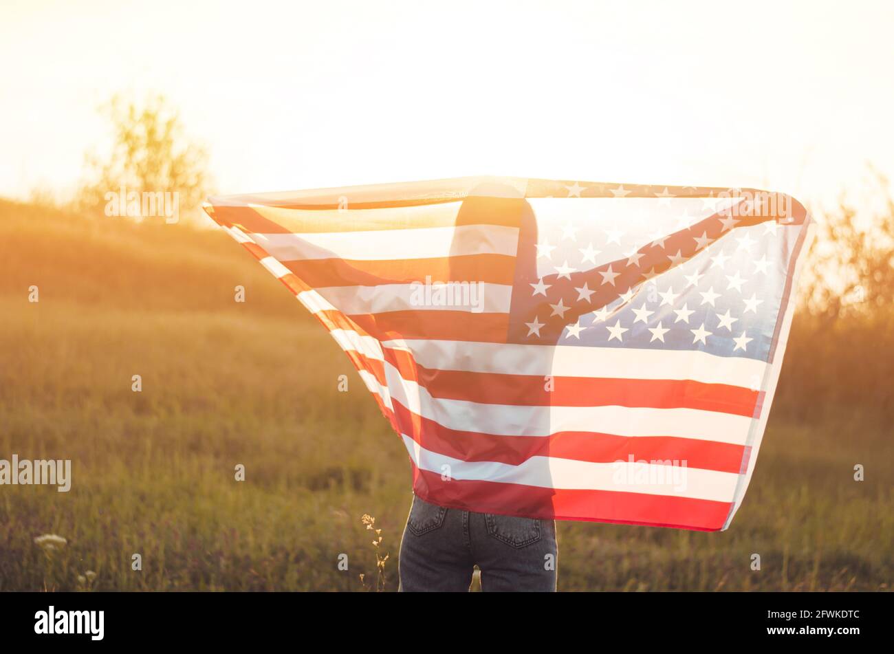 Vue arrière d'une jeune femme portant le drapeau national américain dans un champ au coucher du soleil. Jour de l'indépendance de l'Amérique. Concept de liberté américain Banque D'Images