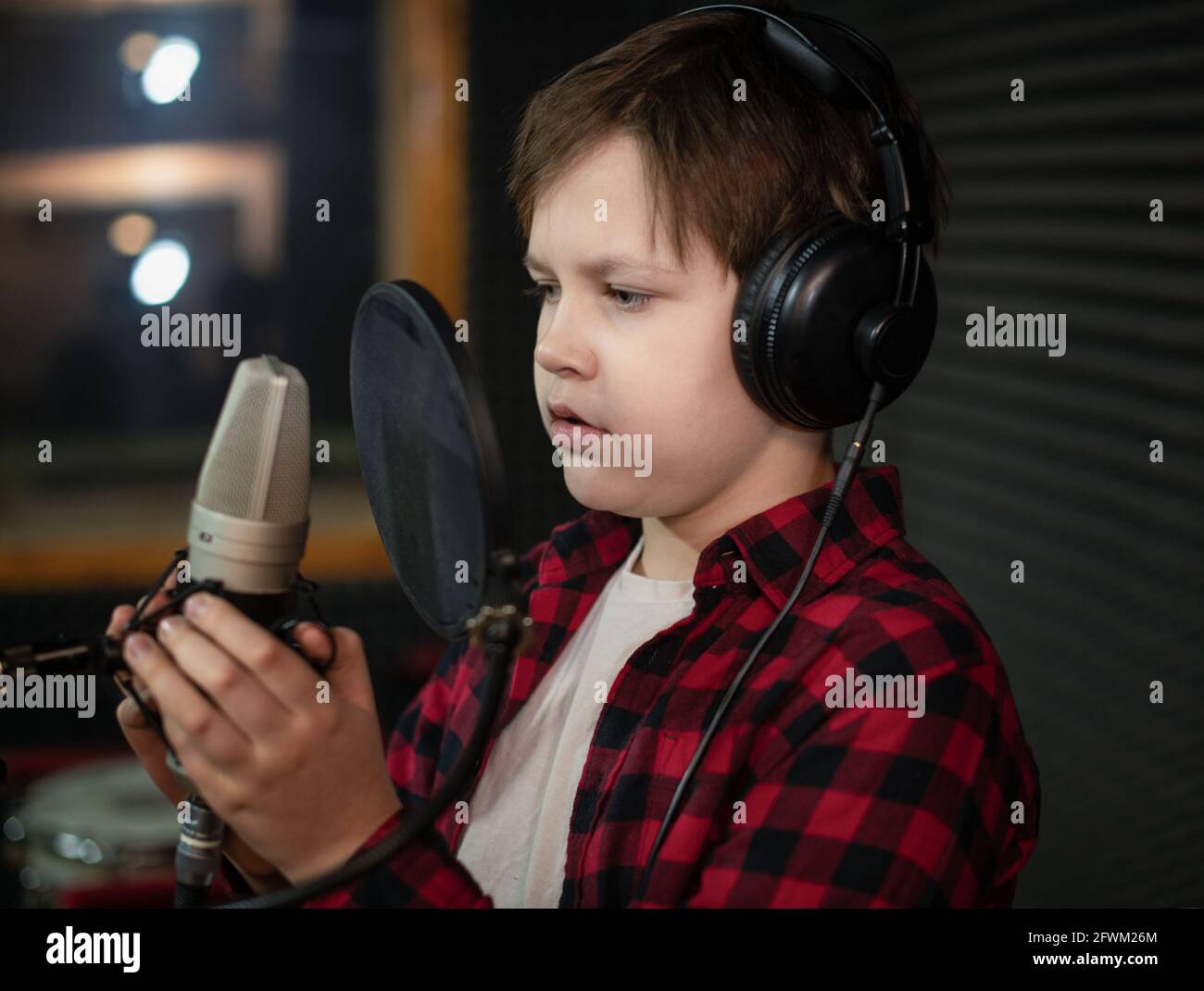 Un garçon dans une chemise à carreaux et un casque chante ou parle dans un microphone avec filtre pop en studio. Banque D'Images