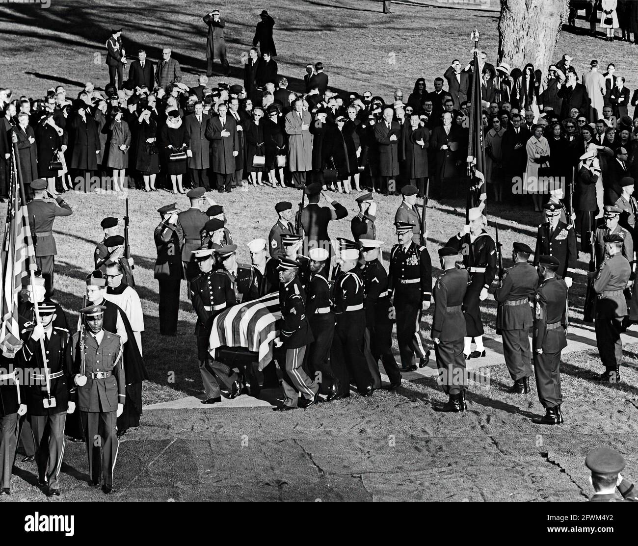 Les pallbearers de la garde d'honneur portent le cercueil à pavillon du président John F. Kennedy sur la tombe du cimetière national d'Arlington, pendant les services de tombes des funérailles d'État du président Kennedy. Marche en procession : archevêque de Washington, Patrick A. O’Boyle; président des chefs d’état-major interarmées, général Maxwell D. Taylor; chef d’état-major de l’Armée de l’air des États-Unis, général Curtis E. LeMay. Les officiels et les dignitaires observent, notamment : le sénateur Kenneth B. Keating (New York); le sénateur Daniel Inouye (Hawaii). Banque D'Images