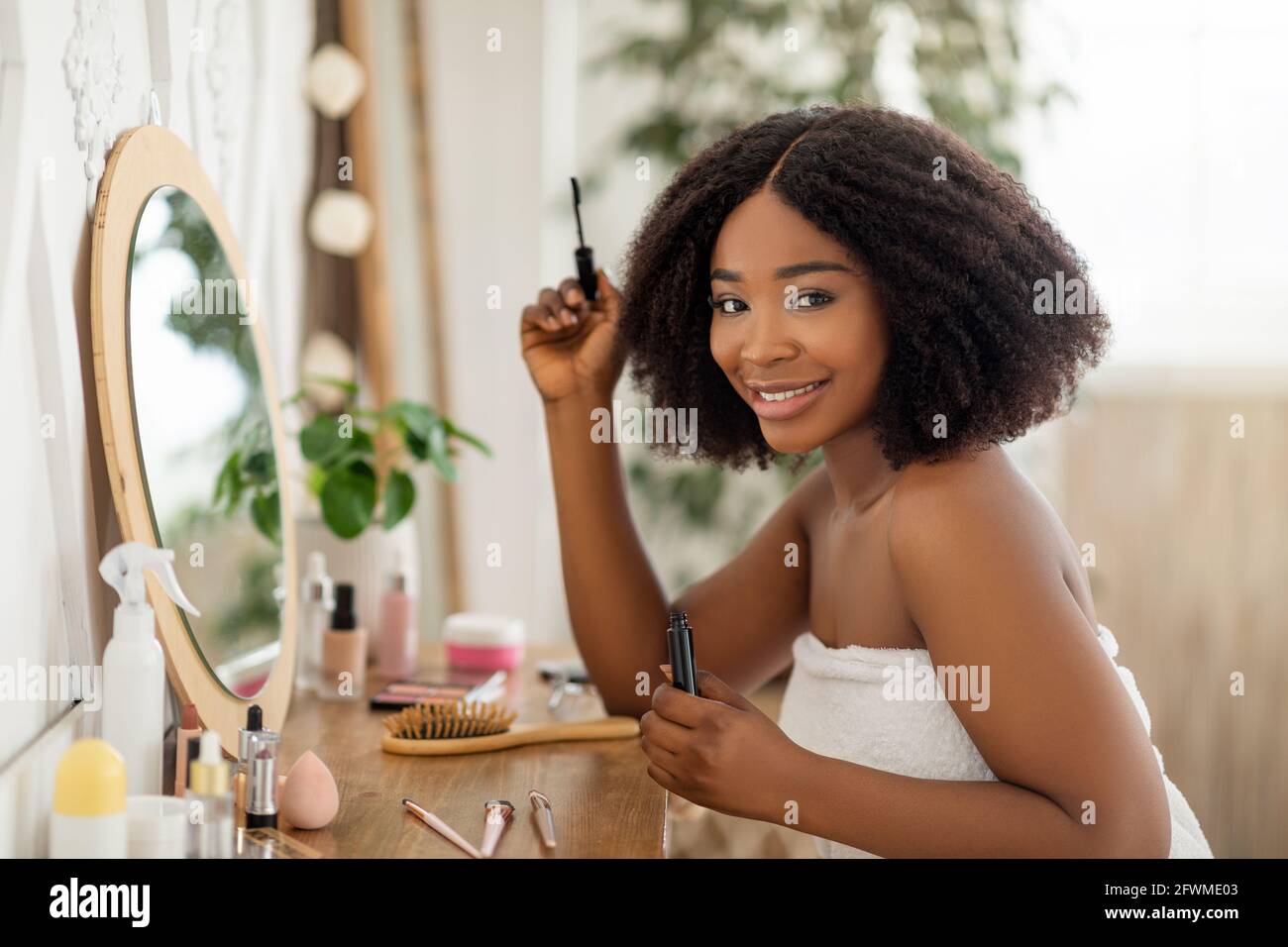 Routine de beauté matinale. Belle dame noire appliquant la mascara, assis à la coiffeuse, regardant dans le miroir à la maison Banque D'Images