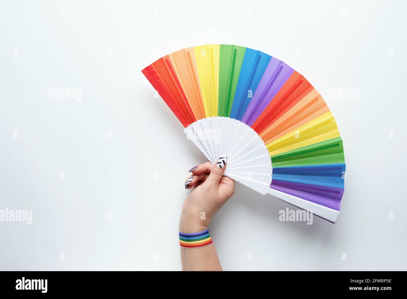 Fierté de la communauté LGBTQ. Mains courantes. Ventilateur rainbow. Ruban avec motif arc-en-ciel au poignet. Pose à plat, vue de dessus sur papier blanc. Simple, minime fierté LGBT Banque D'Images