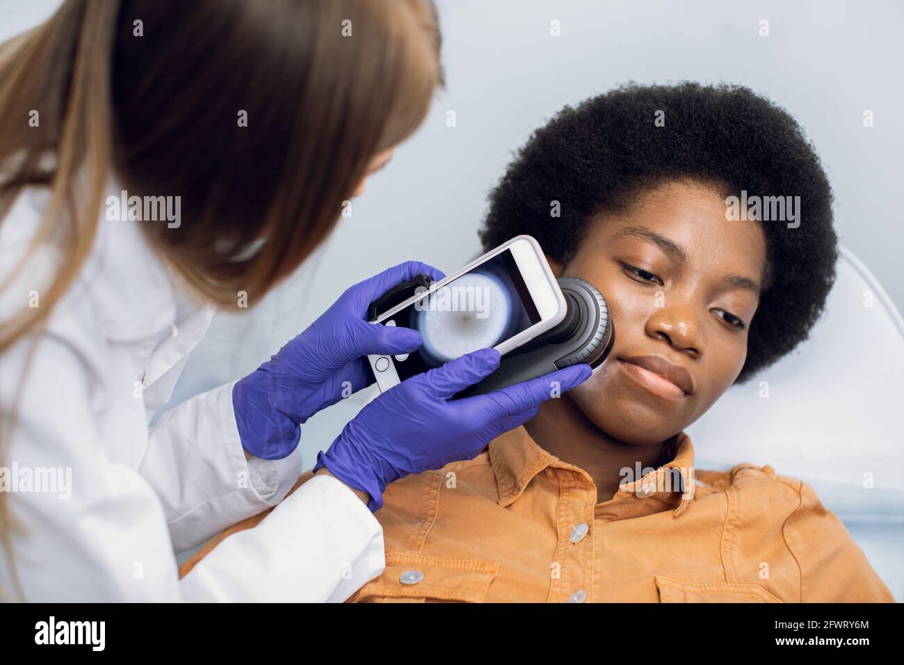 Gros plan des mains de la femme médecin tenant le dermatoscope pour l'étude des moles, des marques de naissance, des verrues, la vérification du visage de sa patiente. Belle afro-américaine couchée sur le canapé Banque D'Images