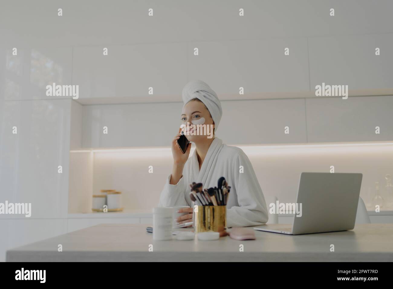 Belle femme en peignoir avec une serviette sur la tête qui parle téléphone mobile lors des procédures de soins de la peau à la maison Banque D'Images