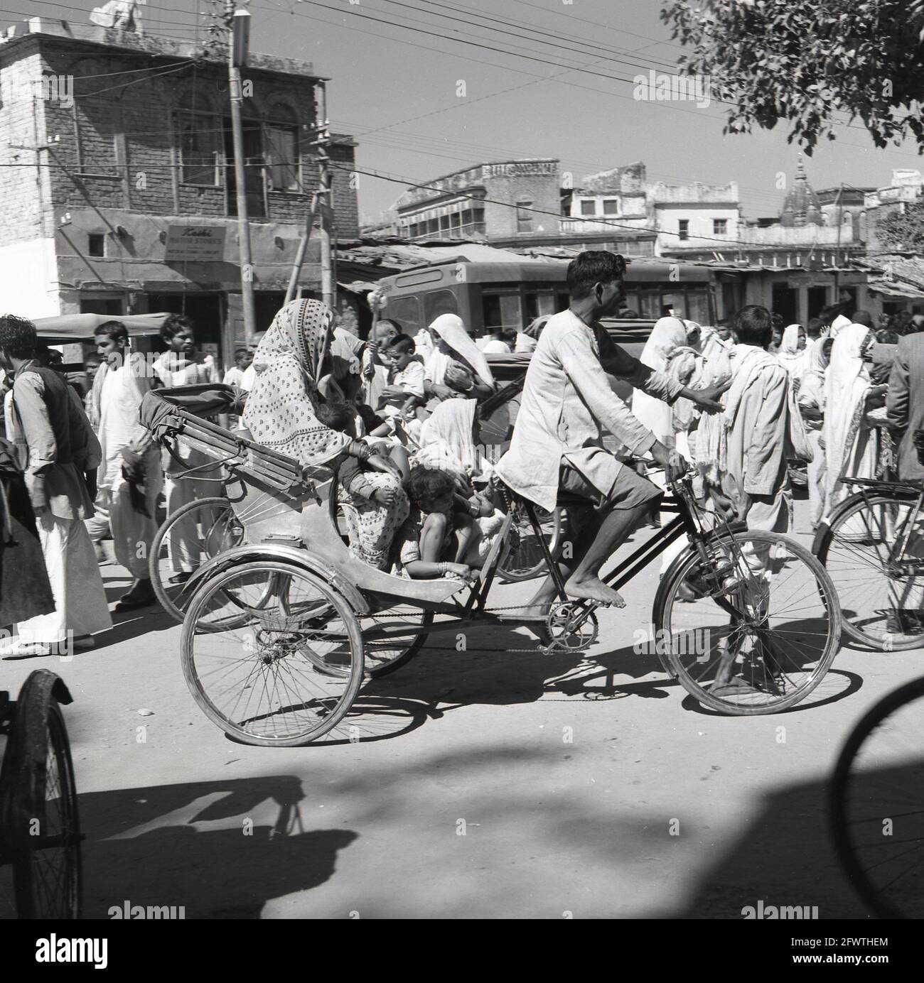 Dans les années 1950, un taxi à vélo, l'Inde, une mère et plusieurs enfants qui se font prendre un tour en pousse-pousse à vélo qui traverse une rue poussiéreuse par un chauffeur de marche en pieds-de-pieds. Ce transport local à petite échelle, une sorte de tricycle à hayon, transporterait des passagers à louer et était donc connu par plusieurs noms : taxi à vélo, taxi à velo, trishaw ou vélo à hayon. Les rickshaws à cycle sont originaires des années 1880. À la fin des années 1920, ils étaient largement utilisés à Singapour et, en 1950, partout dans le sud et l'est de l'Inde. Il y a plusieurs modèles différents, en Inde, les passagers sont assis derrière le conducteur. Banque D'Images