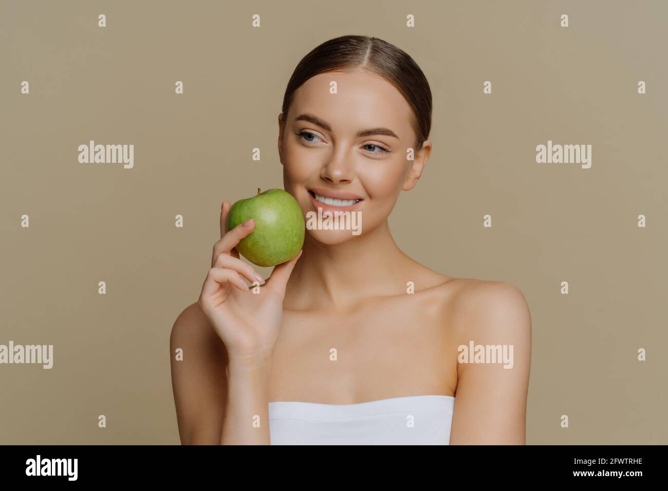 La charmante femme européenne attentionnés tient la pomme près du visage souriant doucement a des dents blanches parfaites saine peau propre enveloppée dans la douche porte-serviettes avec Banque D'Images
