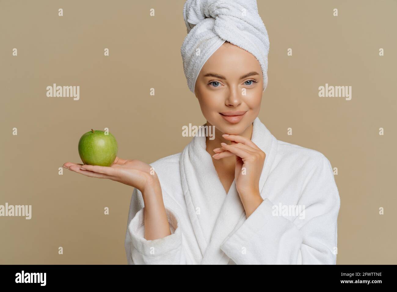Bonne jeune femme européenne a la peau lumineuse parfaite hods la pomme verte fraîche subit des procédures de beauté à la maison habillée vinaigrette blanche pour le bain Banque D'Images