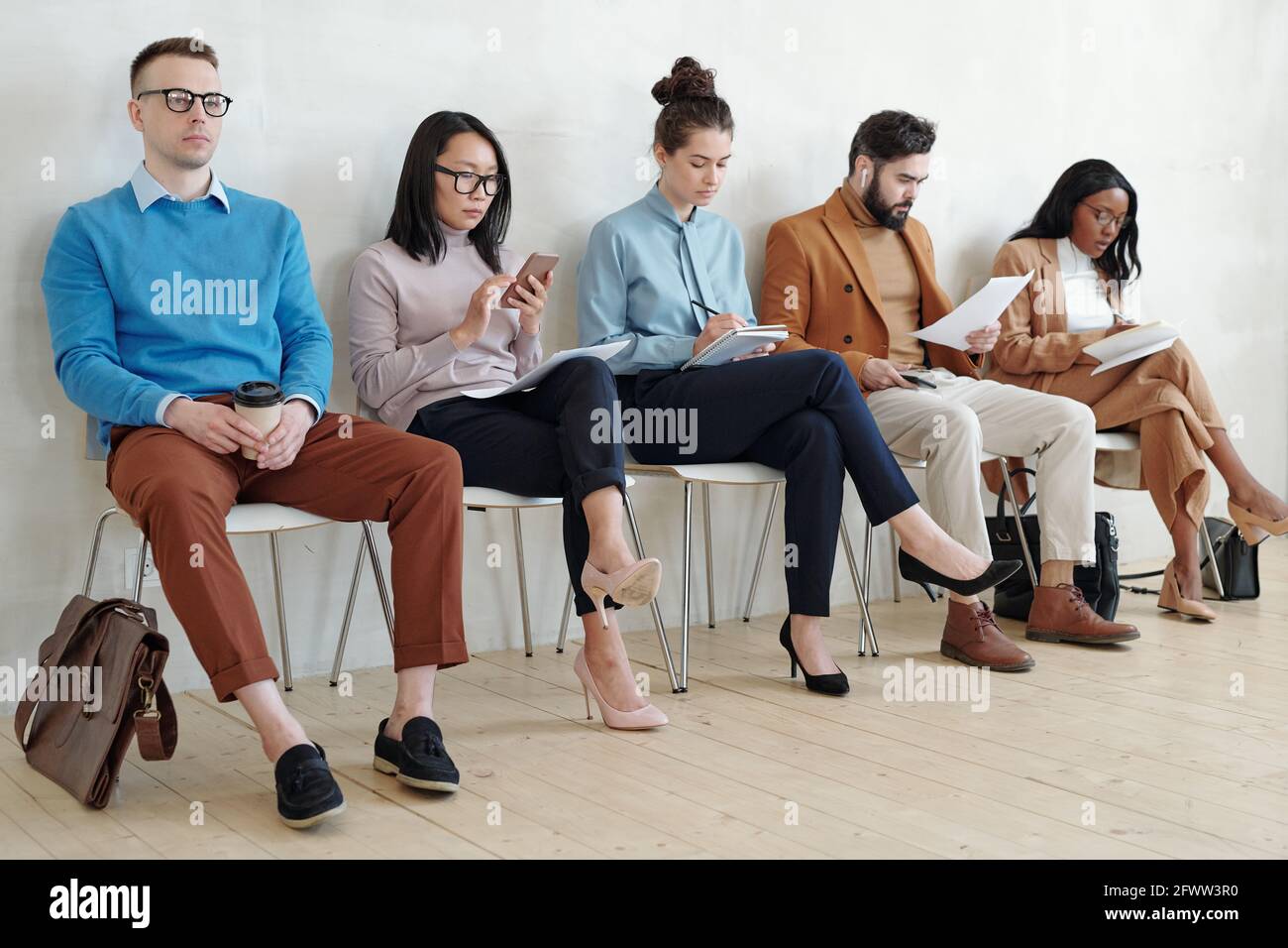 Groupe de jeunes candidats sérieux à un emploi multiethnique dans une ambiance décontractée et intelligente tenues assises sur des chaises en ligne et se préparant à l'entrevue dans un couloir Banque D'Images