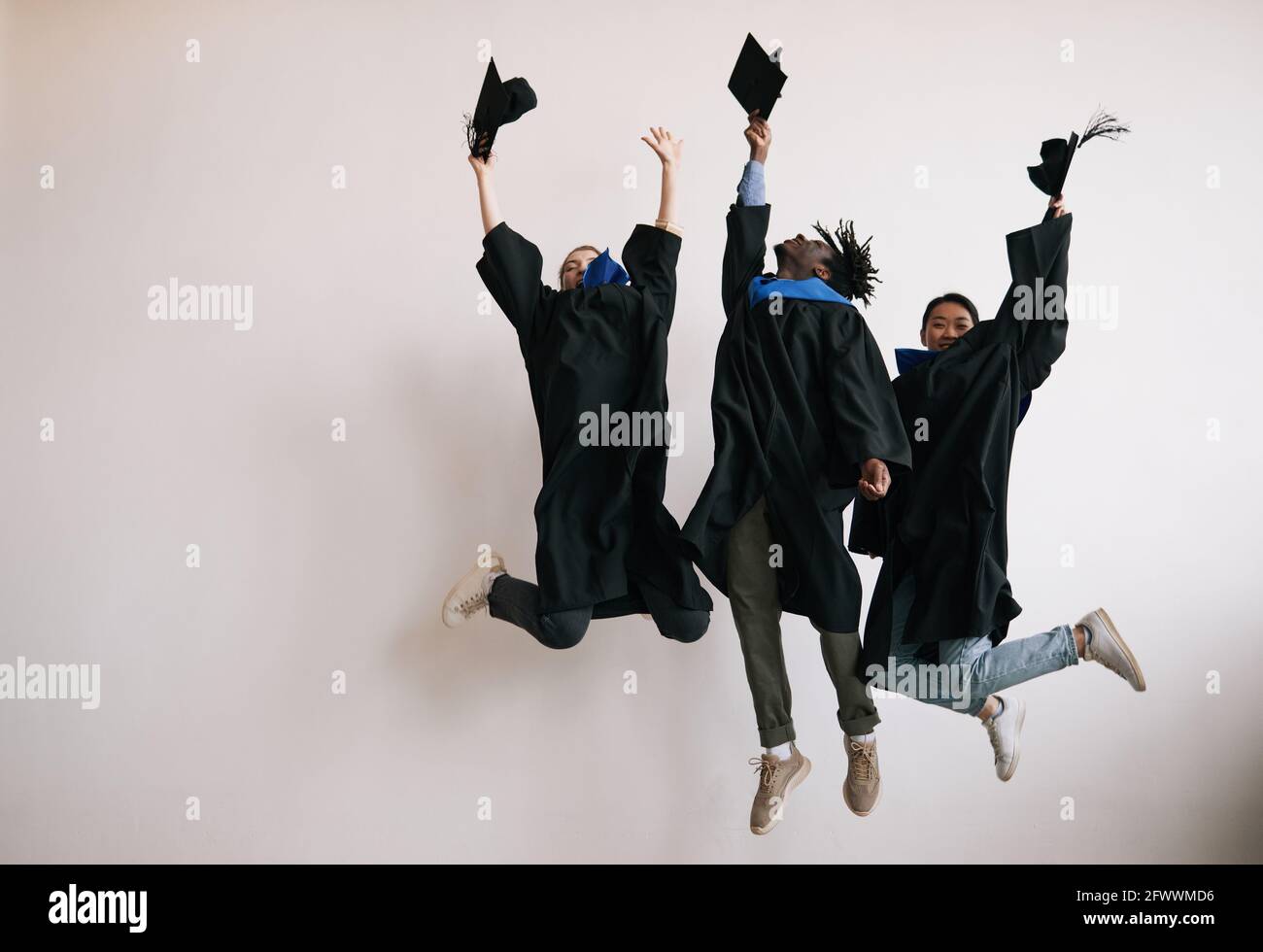 Pleine longueur de trois heureux diplômés d'université portant des robes et sautant sur fond blanc, espace de copie Banque D'Images