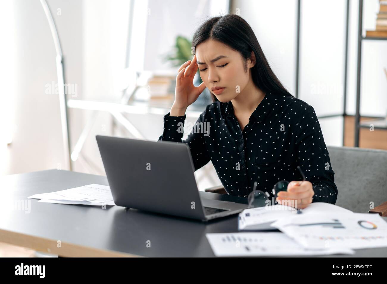 Un travail excessif a bouleversé une femme d'affaires asiatique, une personne indépendante ou une directrice, travaillant sur un ordinateur portable, assis sur son lieu de travail, prenant des pauses, éprouvant du stress, tenant des lunettes dans la main, fermant les yeux Banque D'Images