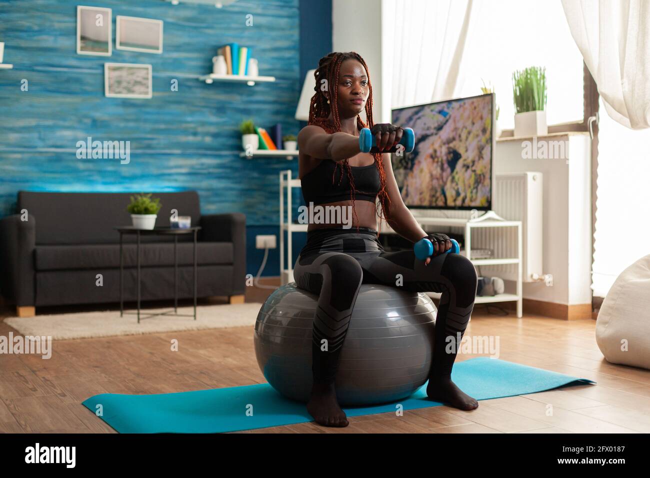 Femme africaine utilisant le ballon de stabilité gardant les bras étirés en train de travailler les épaules à l'aide de haltères bleues, dans la maison salon pour la mise en forme musculaire et un style de vie sain, vêtu de vêtements de sport. Banque D'Images