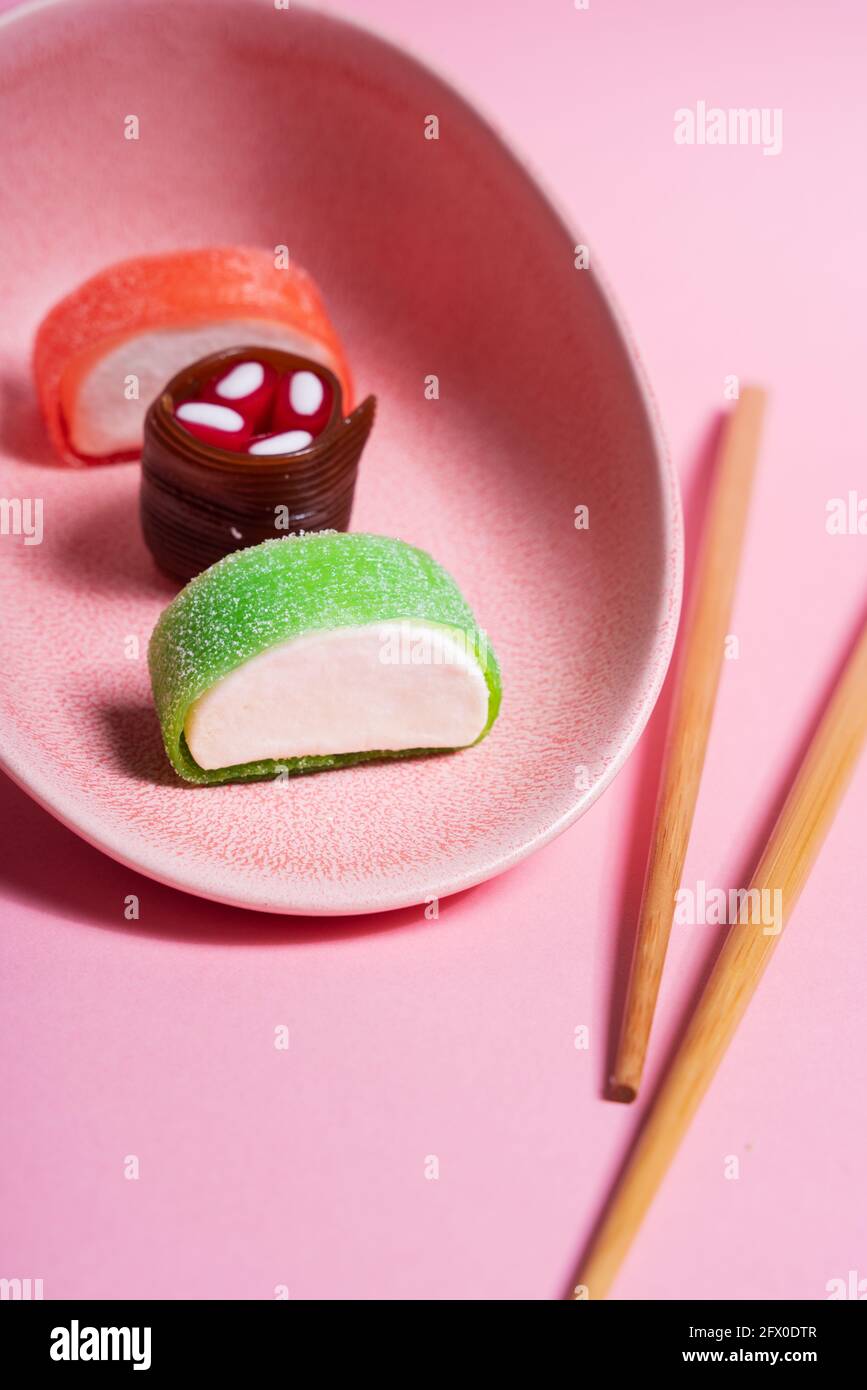 Appétissant sushi doux coloré servi sur une plaque en céramique avec du bois baguettes placées sur fond rose Banque D'Images