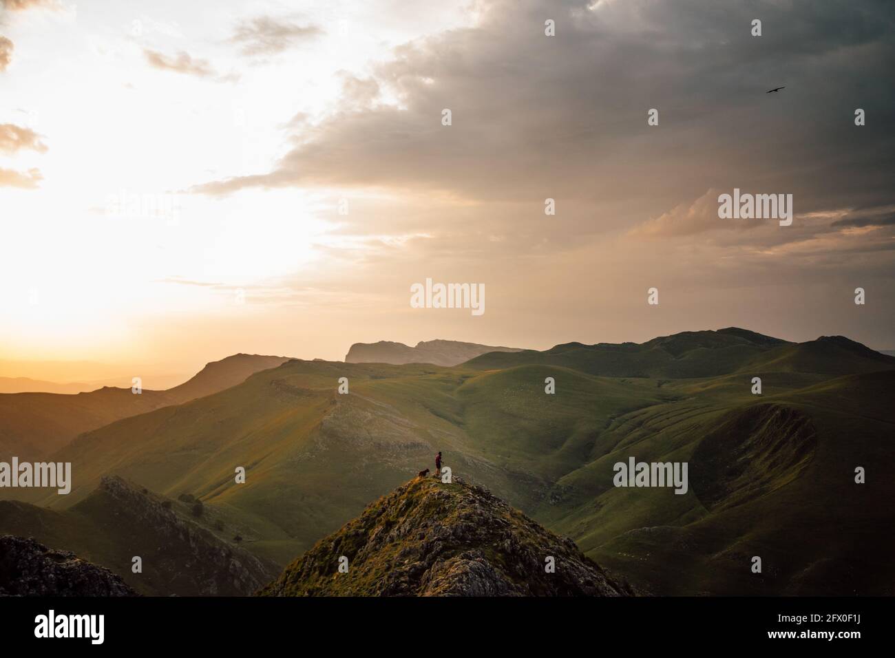 Touriste distant avec chien sur le sommet élevé de la chaîne de montagne avec des pentes dans l'herbe verte sous les nuages Txindoki, Espagne Banque D'Images