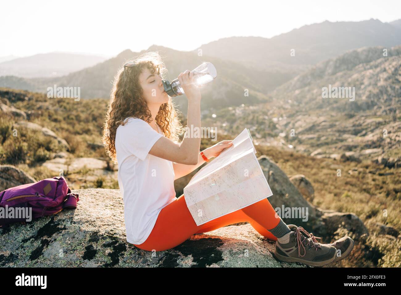 Vue latérale d'une jeune femme en âge de se ranker, avec une obscurité caouate cheveux dans un ensemble décontracté et sac à dos lecture de carte et de boire de l'eau en se reposant sur la pierre pendant Banque D'Images