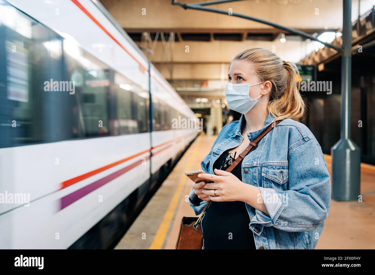 Jeune femme en vêtements décontractés et masque de protection debout gare et vue loin en attendant le train Banque D'Images