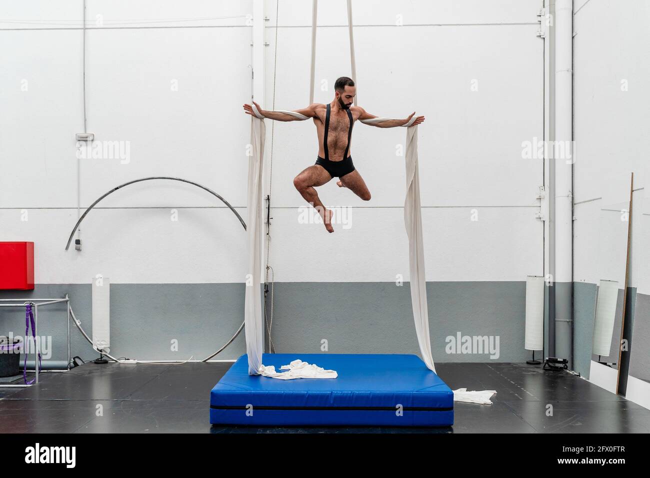 Corps complet musclé sportif robuste en short effectuant de l'exercice sur silks aériens dans un centre de remise en forme moderne et lumineux Banque D'Images