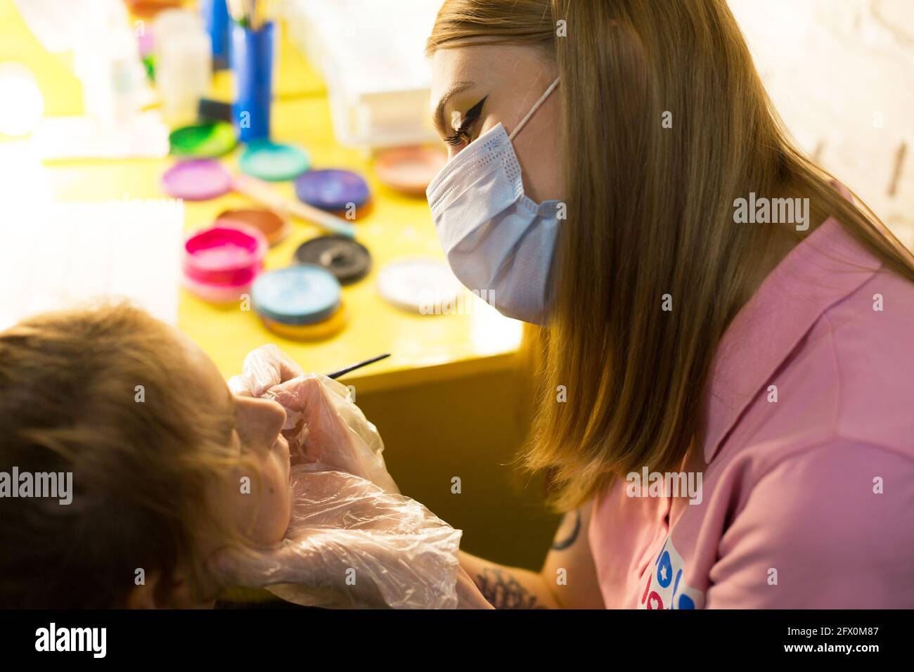 Une femme dans un masque médical dessine un motif aquagrim sur le visage d'un enfant dans le studio devant un miroir avec des lampes. Amusement pour les enfants - coloration du visage. Banque D'Images