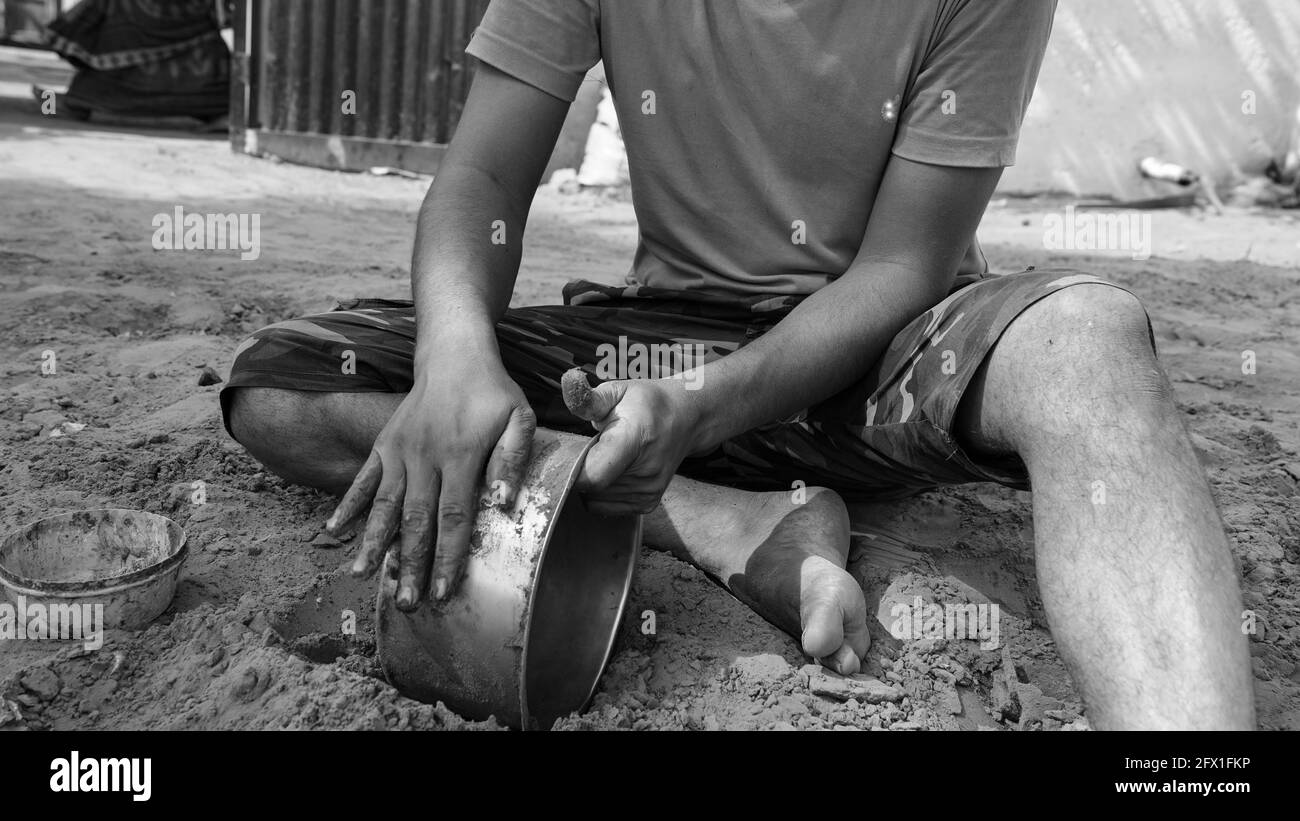 Photo noir et blanc, jeune homme avec chemise rouge essayant de nettoyer un pot en laiton avec de la terre et de l'eau. Le sol pur est le meilleur antibactérien pour nettoyer les pots. Banque D'Images