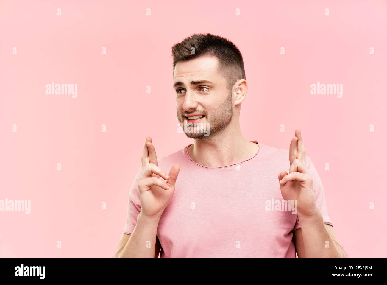 Jeune homme beau croisant ses doigts et souhaitant bonne chance sur fond rose. Concept espoir et souhait Banque D'Images