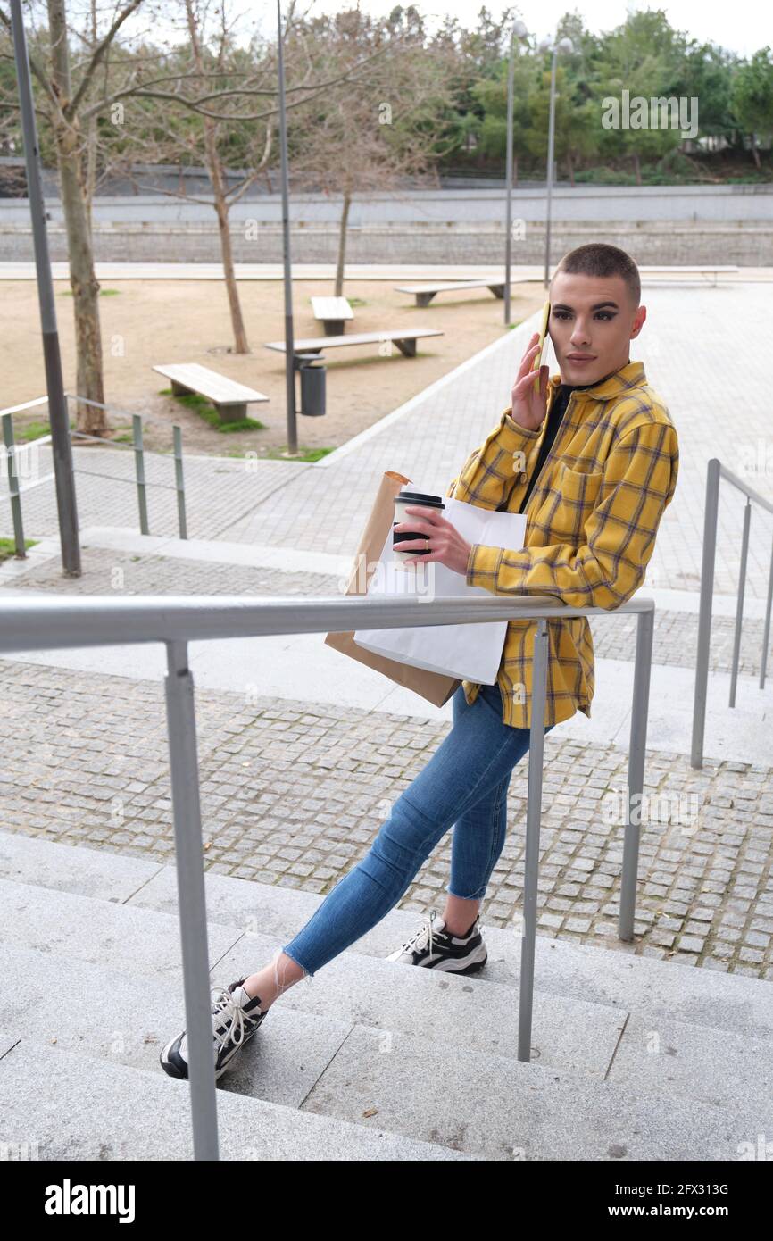 Jeune personne non binaire parlant sur le smartphone avec des sacs d'achats et une tasse de café. Faites le maquillage de la personne androgyne. Banque D'Images