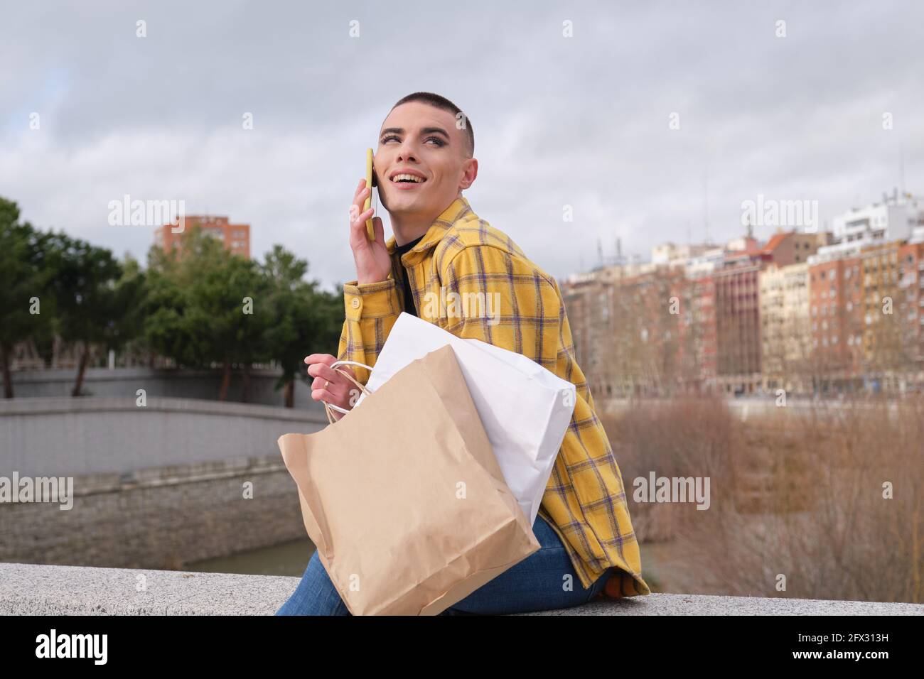 Jeune personne non binaire parlant et riant sur le smartphone avec des sacs d'achats assis sur un mur. Faites le maquillage de la personne androgyne. Banque D'Images