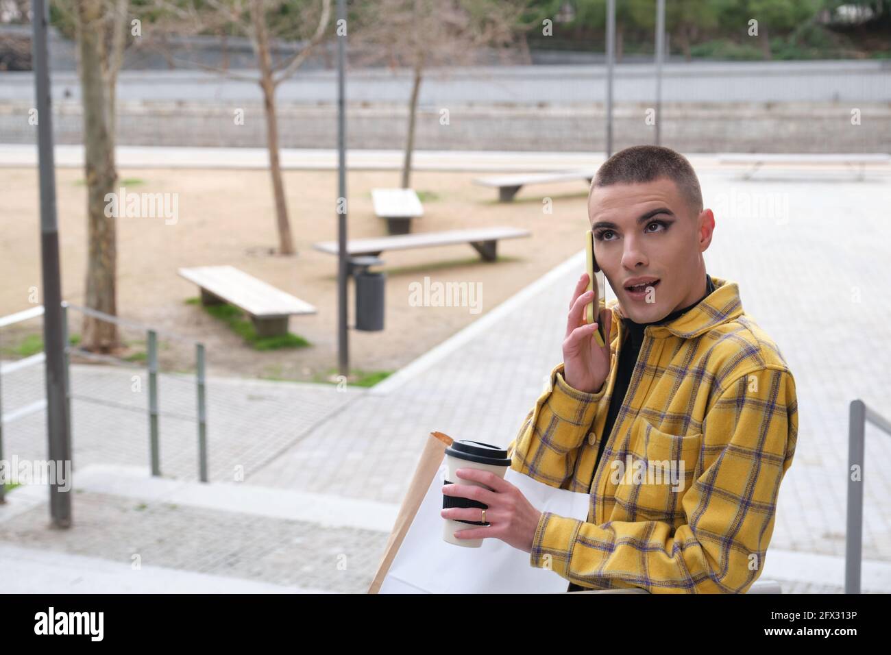 Jeune personne non binaire souriant et parlant sur le smartphone avec des sacs d'achats et une tasse de café. Faites le maquillage de la personne androgyne. Banque D'Images