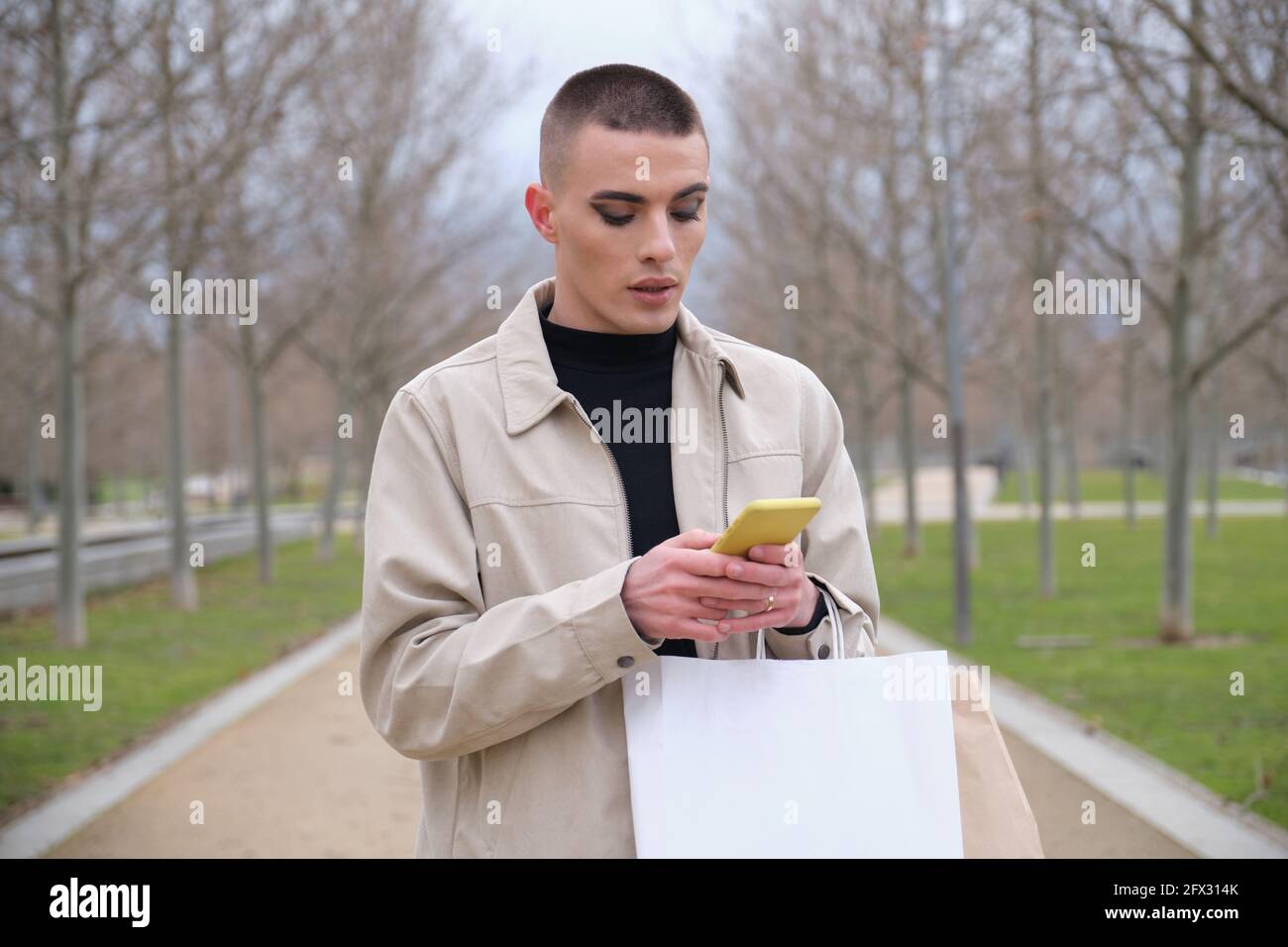 Jeune homme portant un maquillage tenant des sacs de shopping et bavardant sur un smartphone dans un parc. Queer personne. Banque D'Images