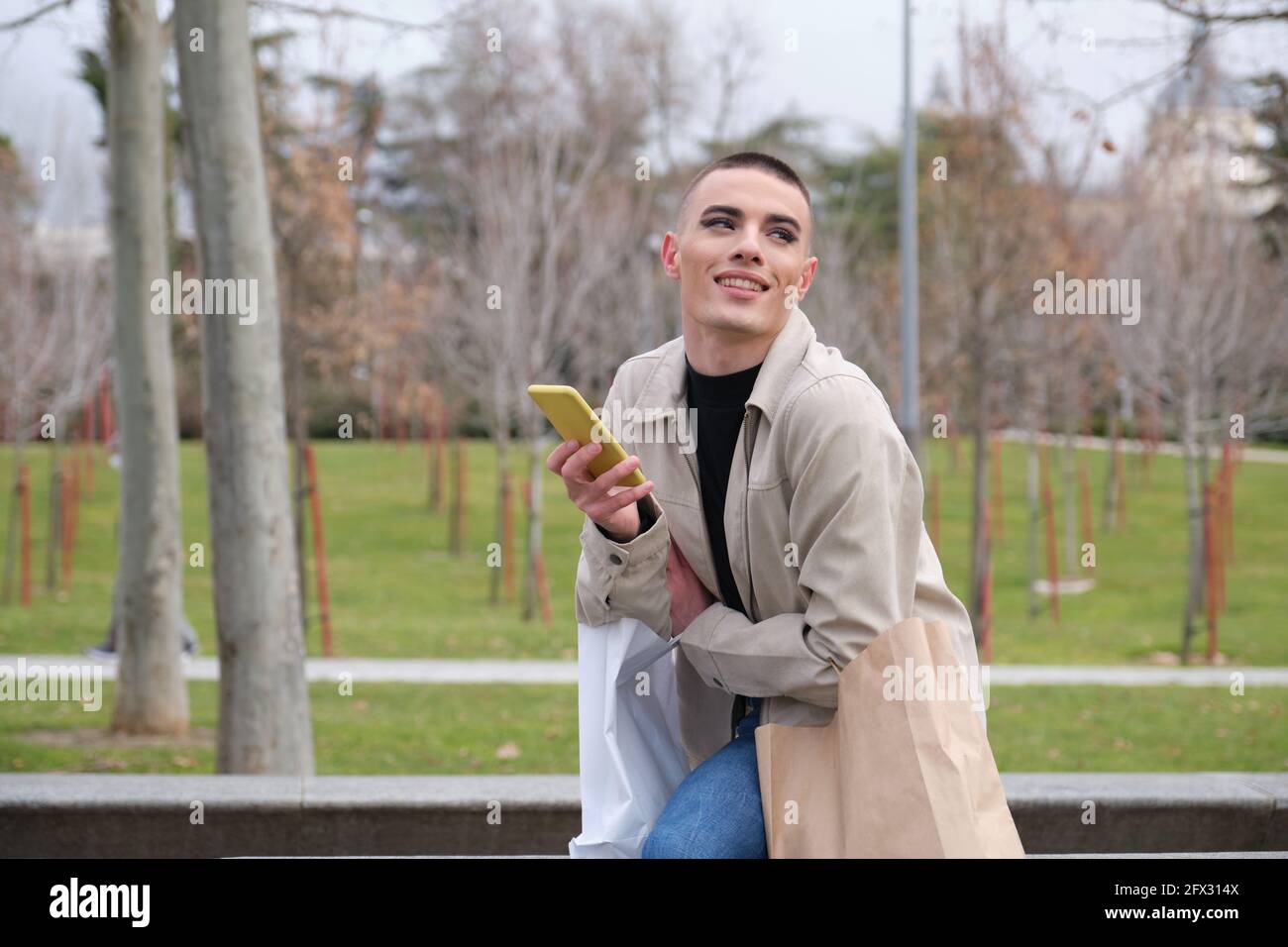 Jeune homme portant du maquillage tenant des sacs de shopping et un smartphone riant assis dans un parc. Queer personne. Banque D'Images