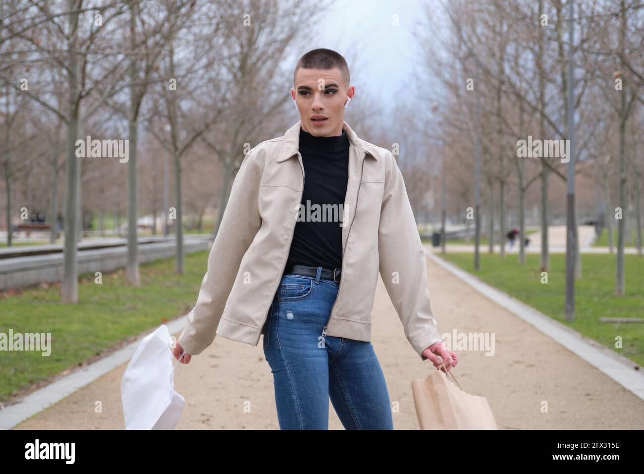 Un jeune homme portant du maquillage tourne autour de sacs de shopping et écoute de la musique dans un parc. Queer personne. Banque D'Images
