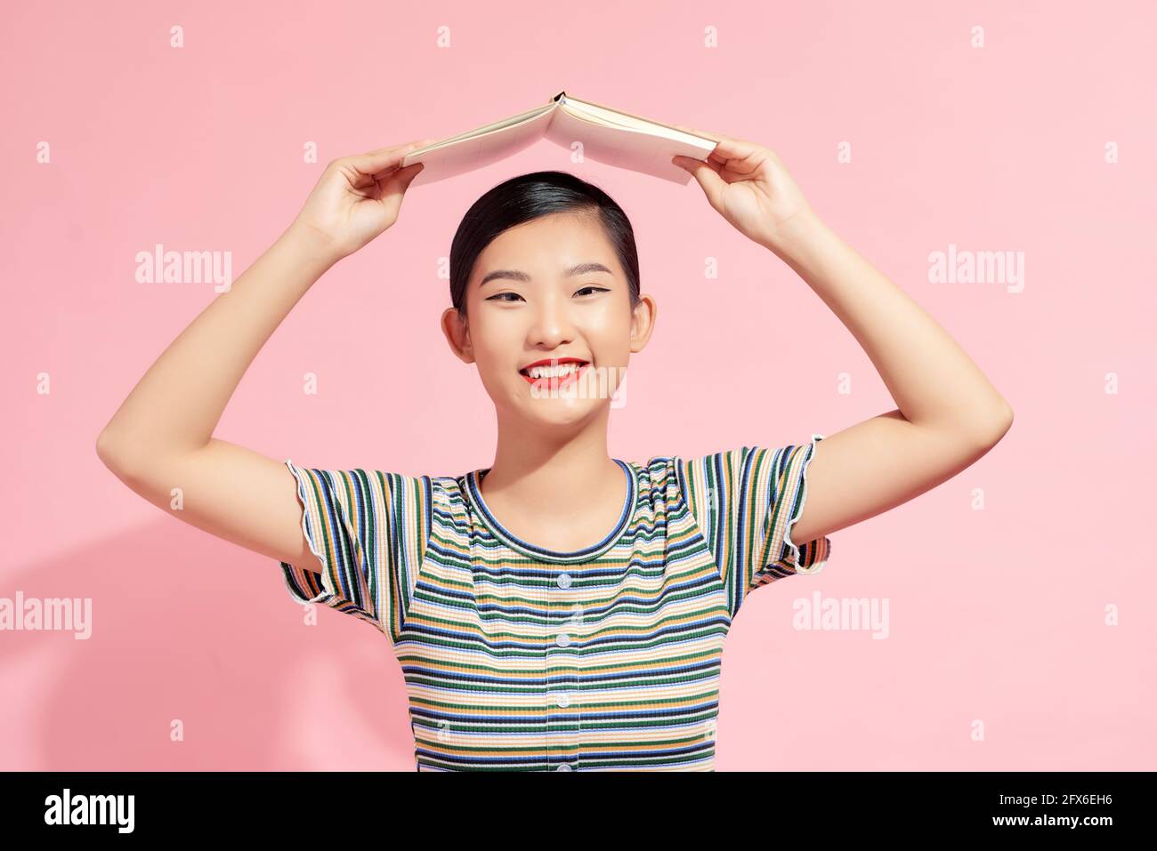 Portrait d'une jeune fille étudiante drôle et souriante avec un ouvert livre sur sa tête sur fond rose Banque D'Images