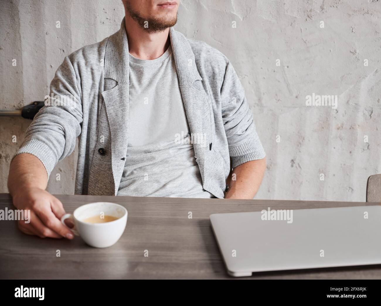 Vue rognée d'un homme reposant avec une tasse de café après le travail sur un ordinateur portable. Le jeune homme en chandail gris est assis au bureau et relaxant. Concept de frein à café. Banque D'Images