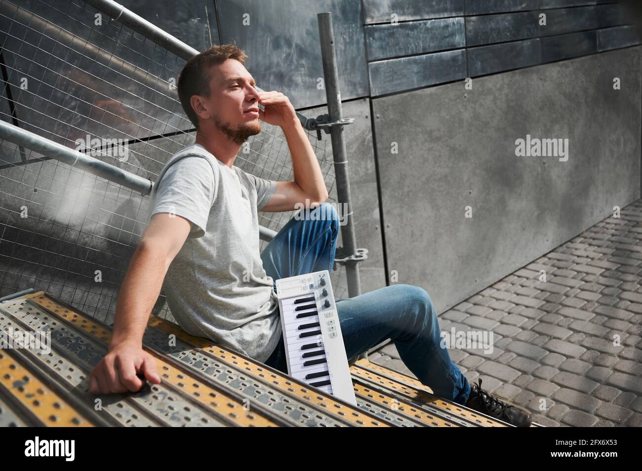 Vue latérale de l'homme avec clavier de piano est assis sur les escaliers. Le jeune homme en T-shirt gris se repose sur les escaliers de la rue et bénéficie du temps ensoleillé. Concept de temps libre. Banque D'Images