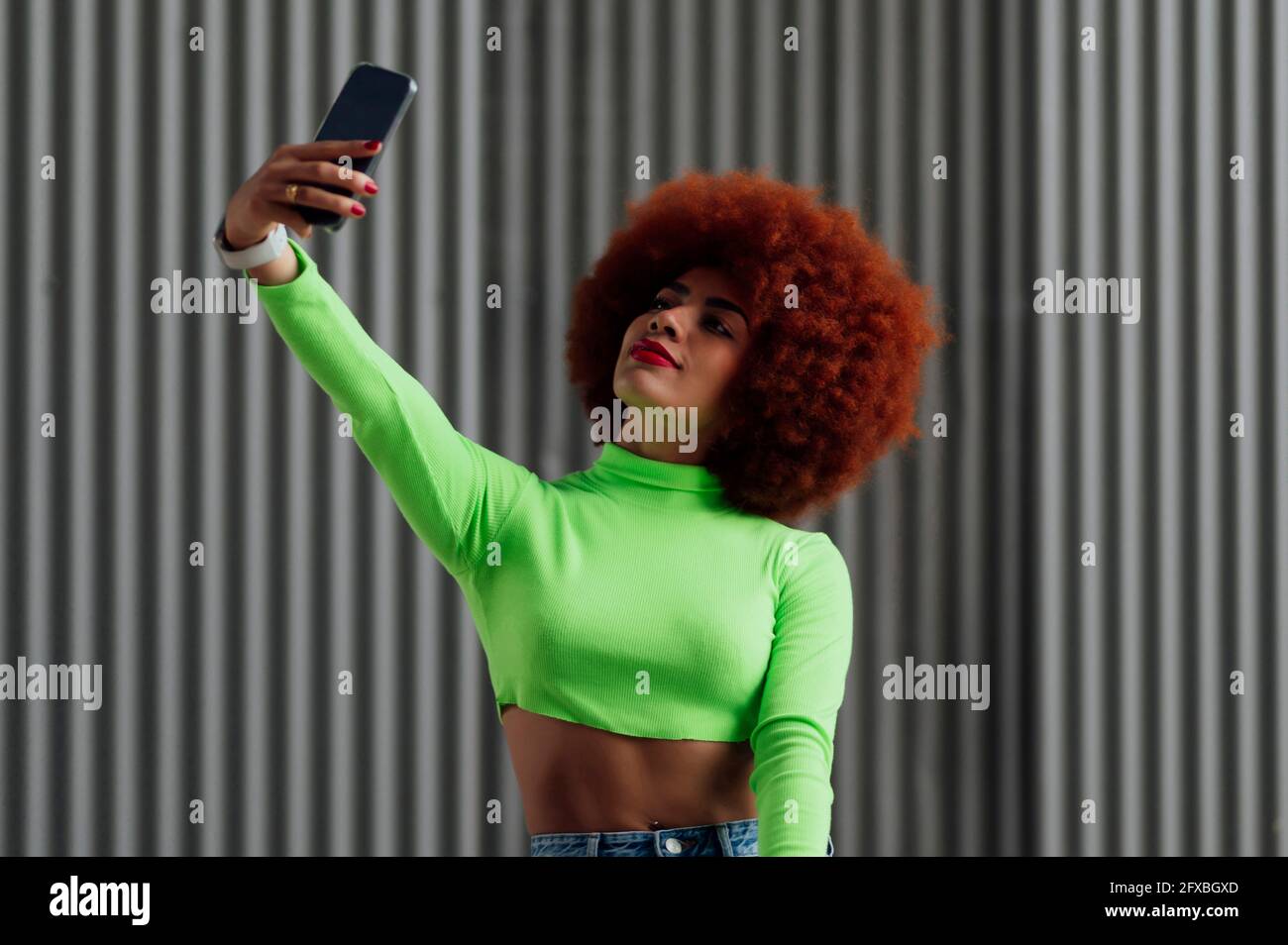 Femme à tête rouge prenant le selfie à travers le téléphone portable devant le mur gris Banque D'Images