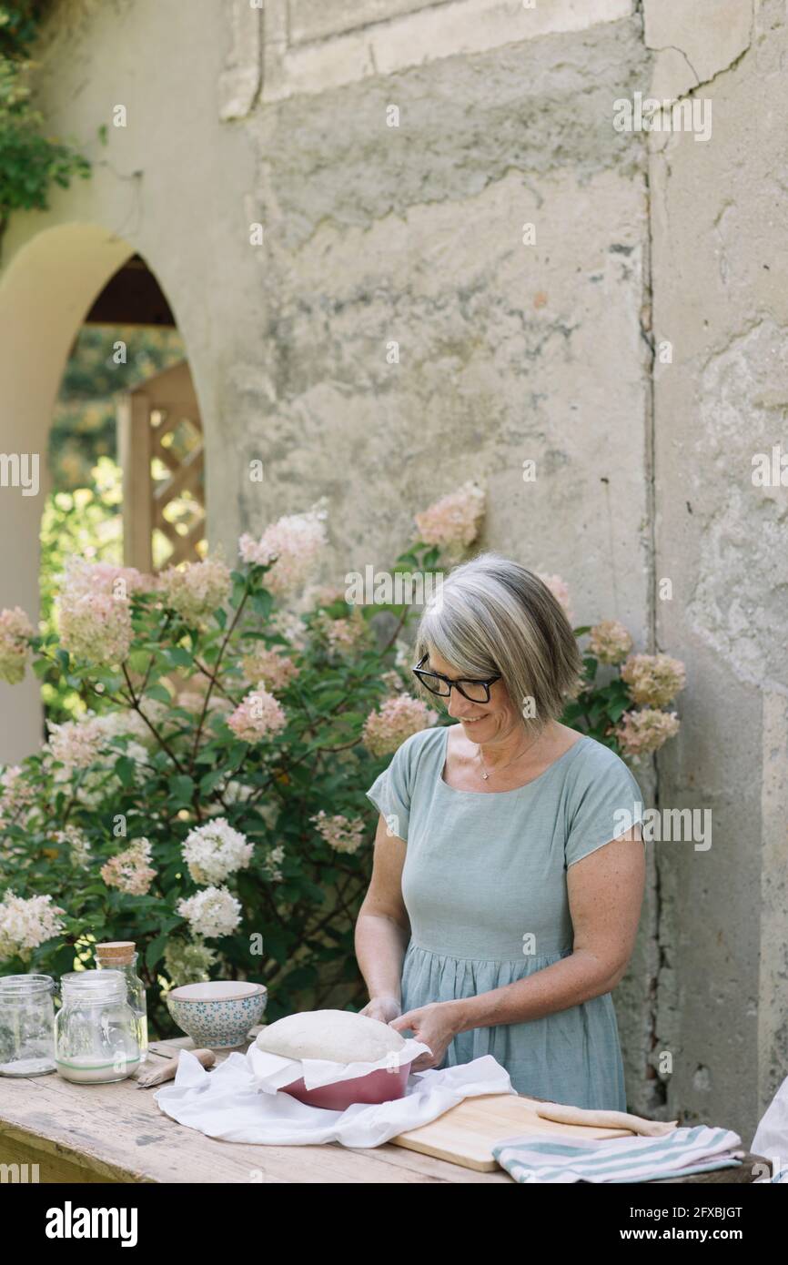 Femme mûre souriante debout près de la plante de fleur tout en préparant le pain à la cour arrière Banque D'Images