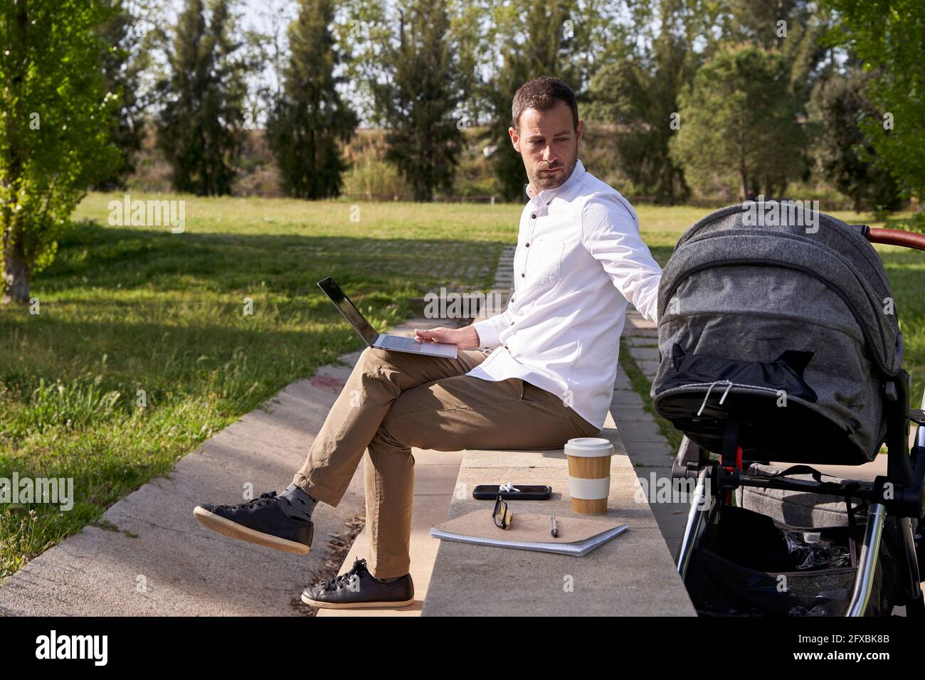 Père regardant la poussette de bébé tout en étant assis avec un ordinateur portable au parc Banque D'Images