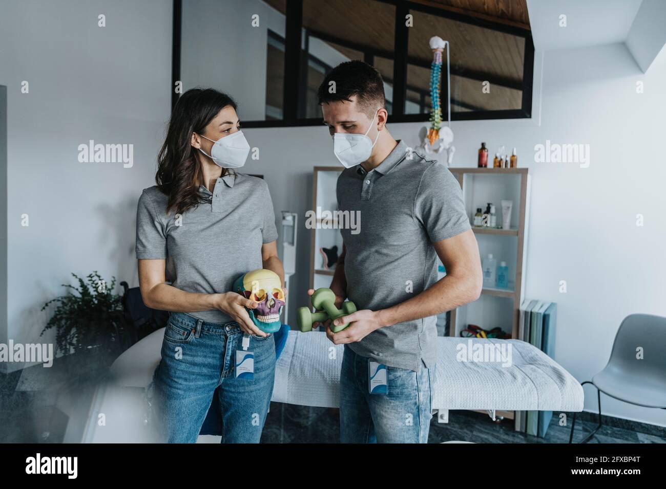Thérapeute physique féminine avec un objet crâne debout collègue mâle tenant des poids devant la table de massage en pratique pendant une pandémie Banque D'Images