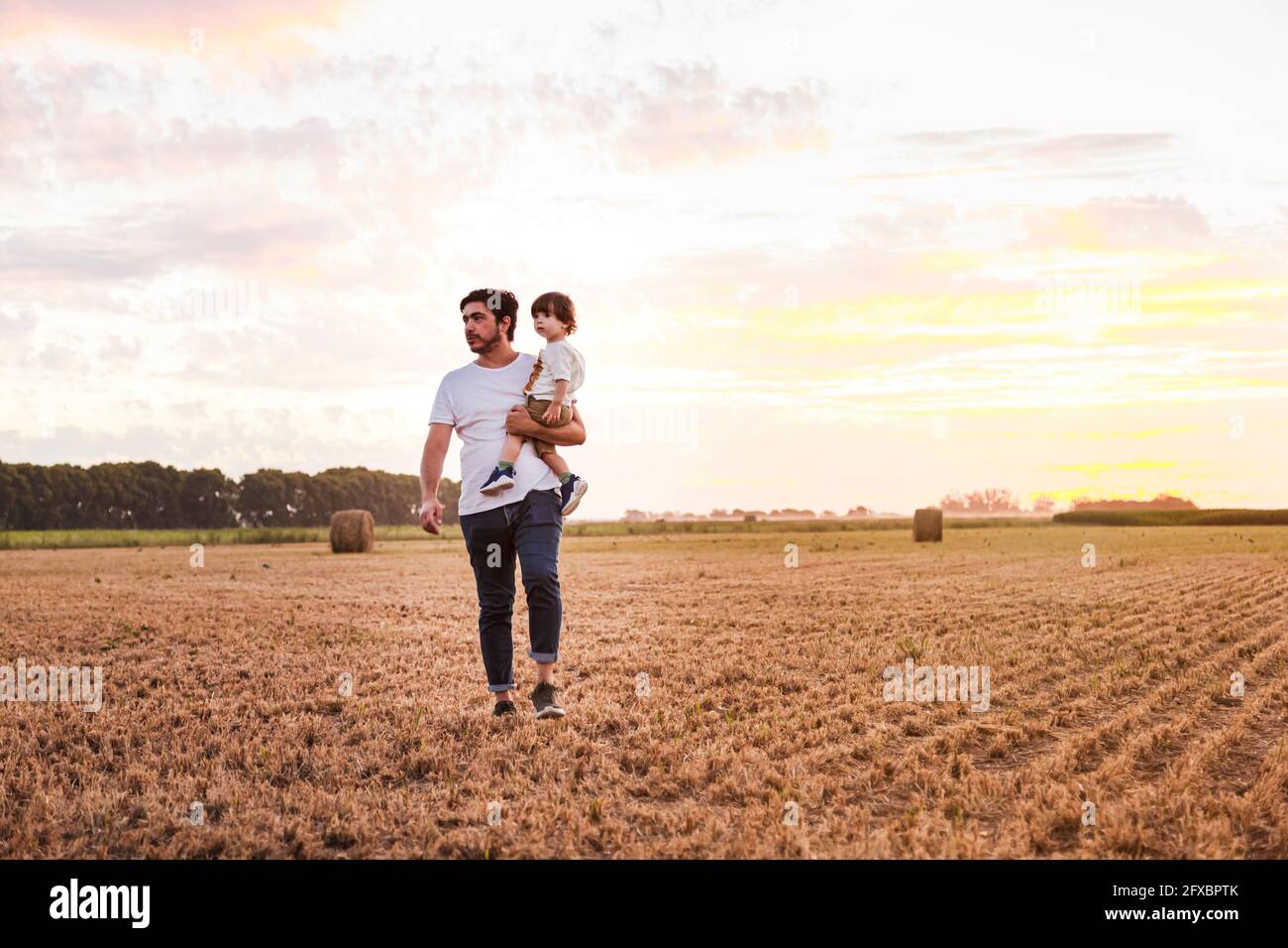 Père transportant son bébé à travers le champ récolté au coucher du soleil Banque D'Images