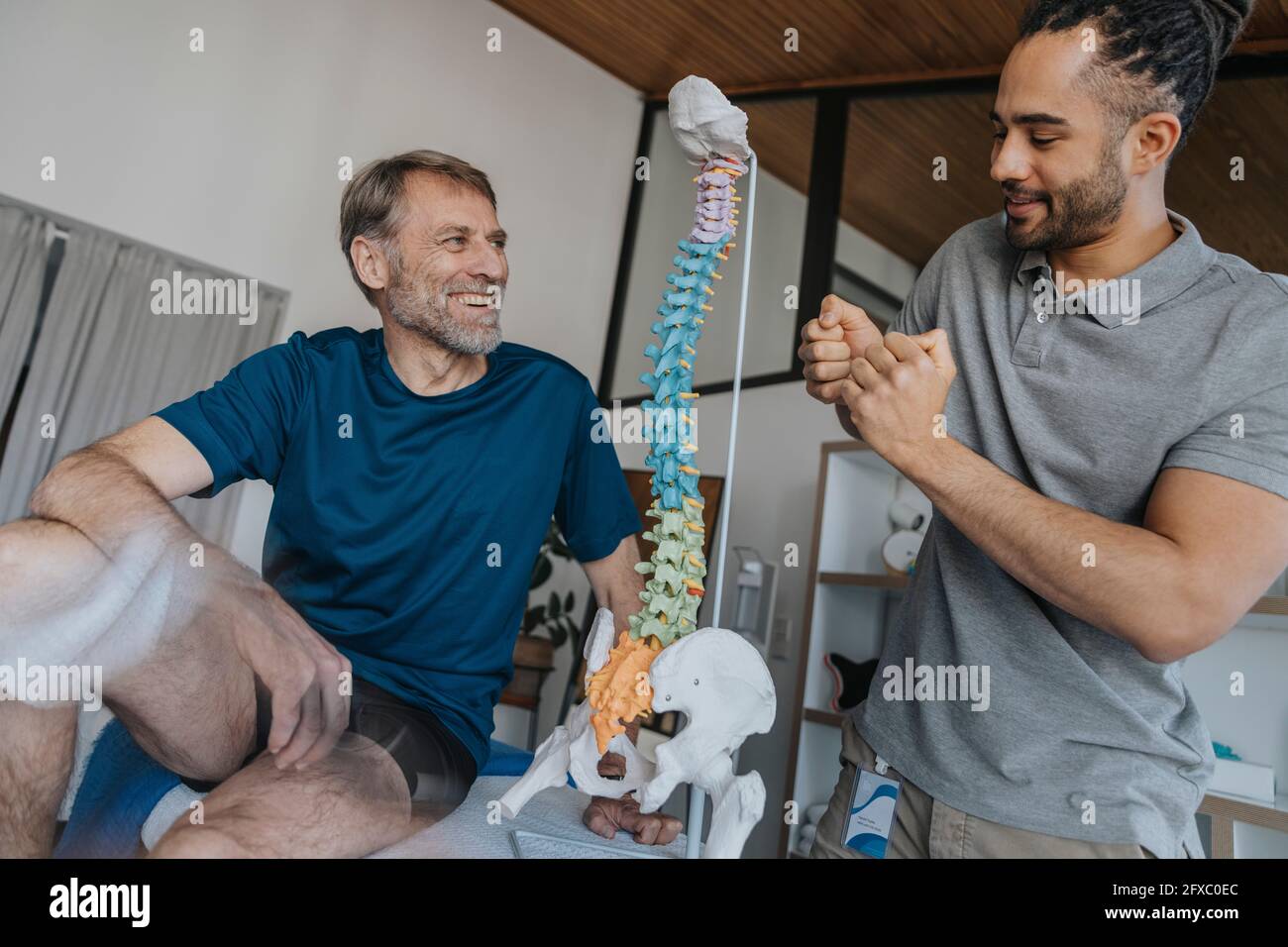 Sourire un homme regardant le physiothérapeute gestant par le modèle de colonne vertébrale dans la pratique Banque D'Images