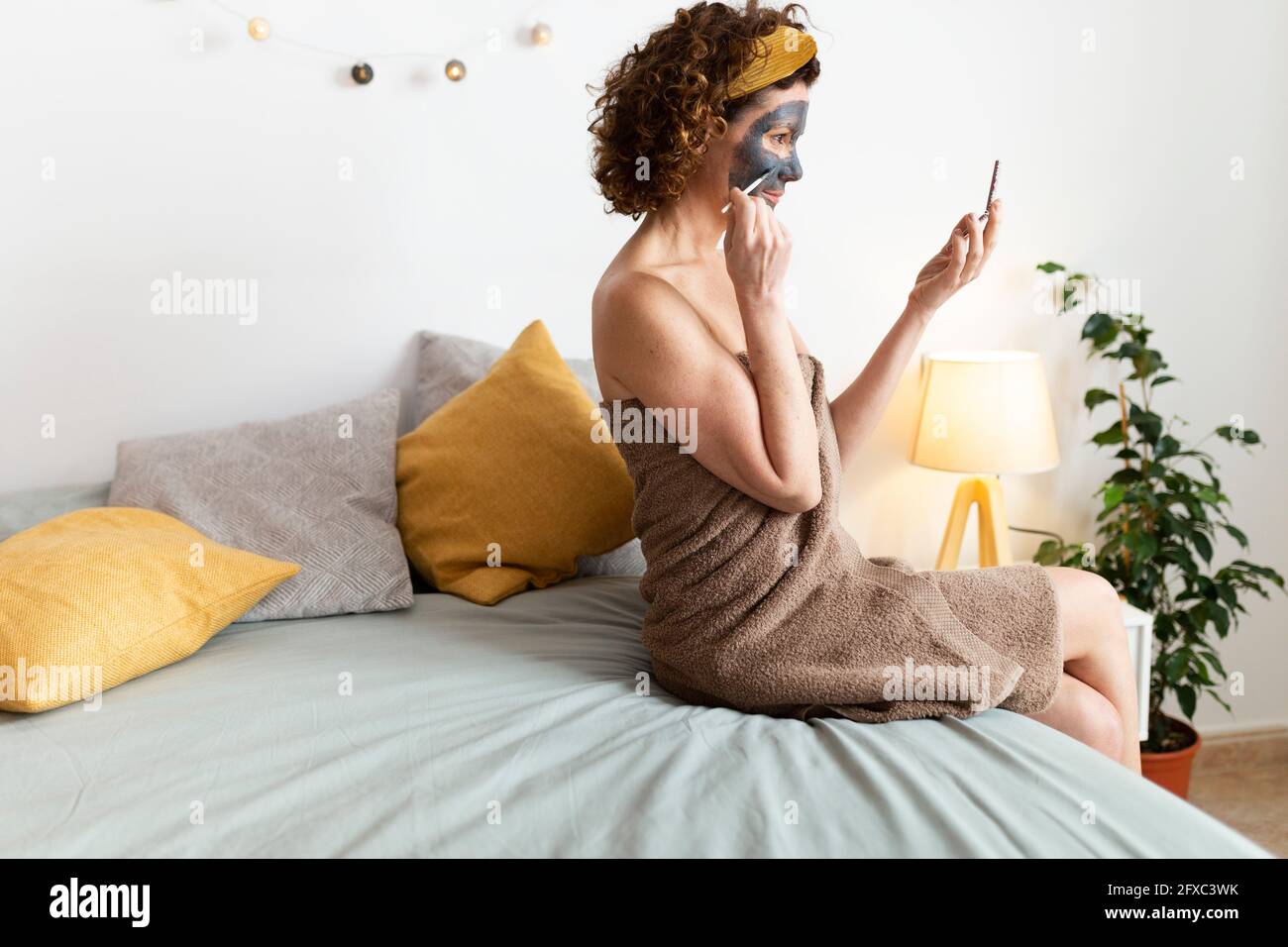 Femme appliquant un masque facial assis sur le lit à la maison Banque D'Images