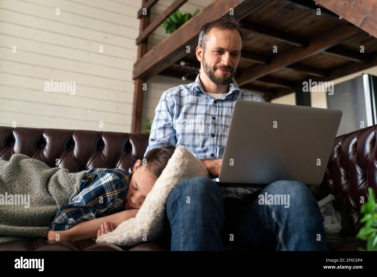 Homme utilisant un ordinateur portable avec son fils dormant sur un canapé à la maison Banque D'Images