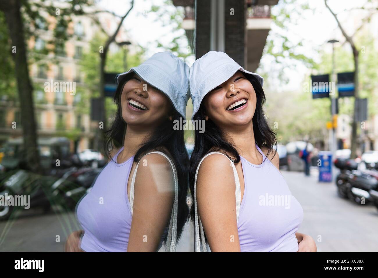 Femme souriante avec chapeau de seau incliné sur le mur de verre dans la rue Banque D'Images