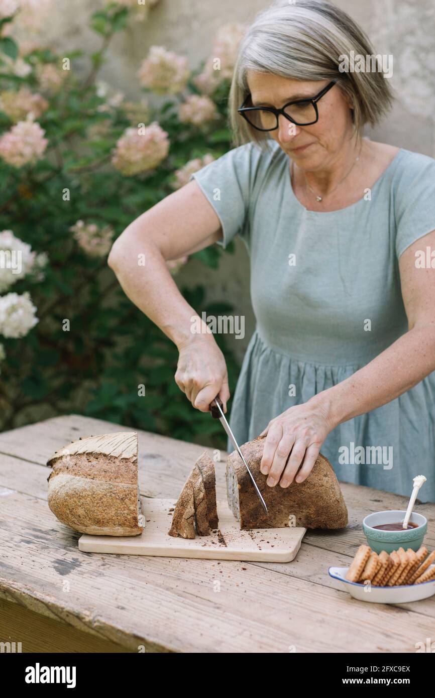 Femme mûre portant des lunettes coupant du pain sur une table Banque D'Images