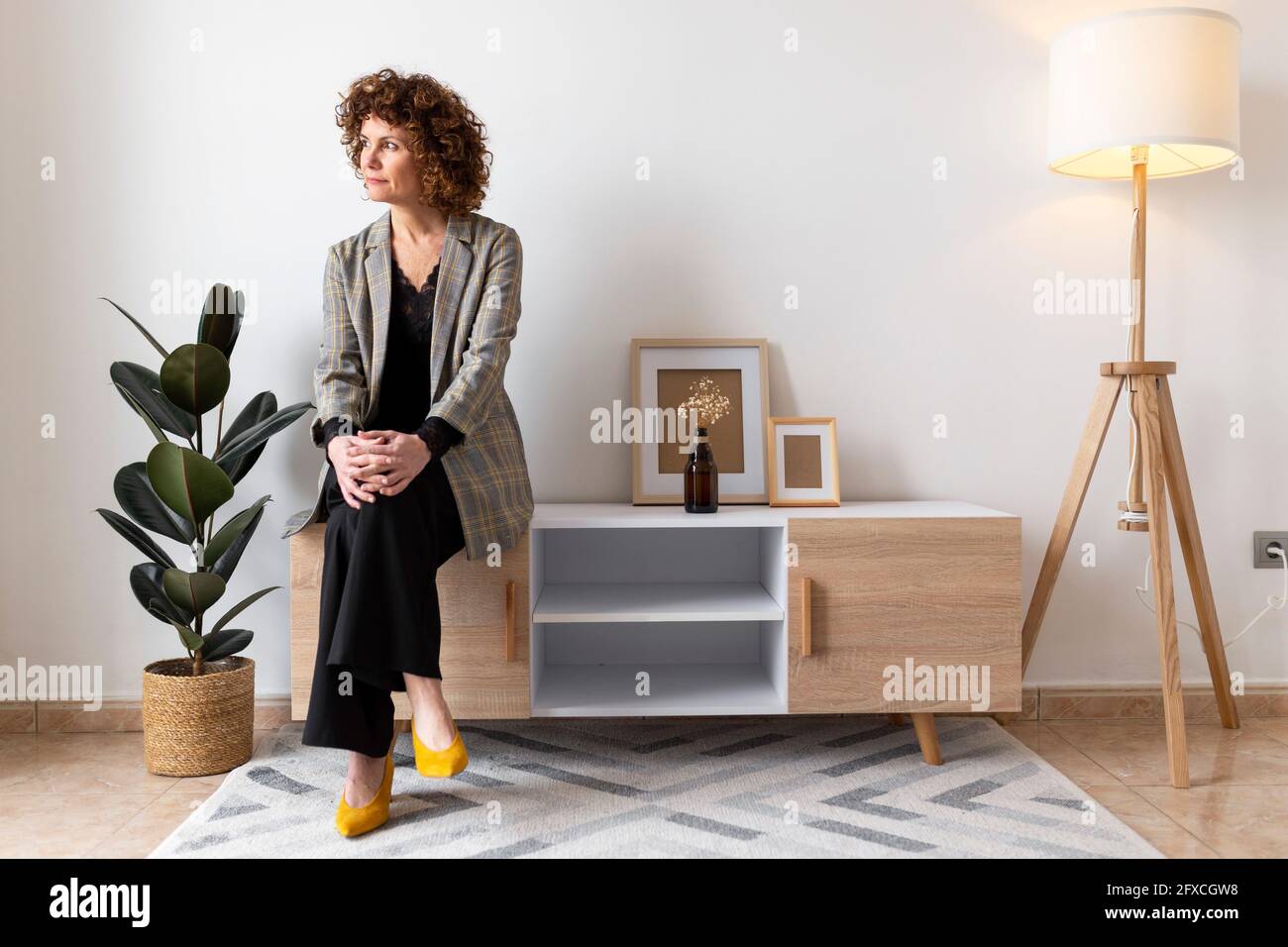 Femme mûre regardant loin tout en étant assise sur un meuble en bois à la maison Banque D'Images