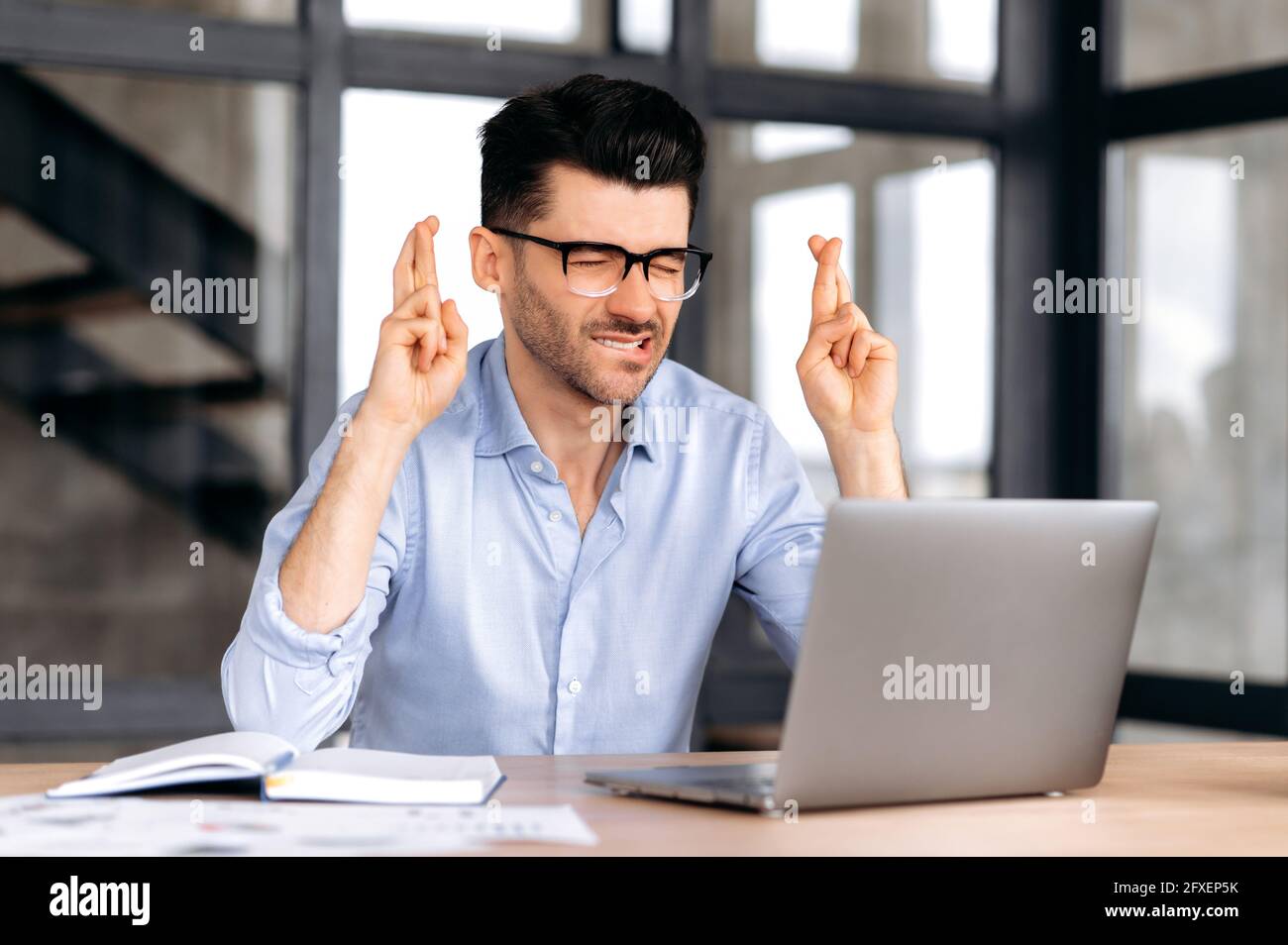 Inquiet caucasiens courtier, Manager ou indépendant portant des lunettes, assis au travail au bureau, en utilisant un ordinateur portable, en espérant de bonne chance, en souhaitant le succès, en se faisant des gestes avec ses mains, ferme les yeux, priant Banque D'Images