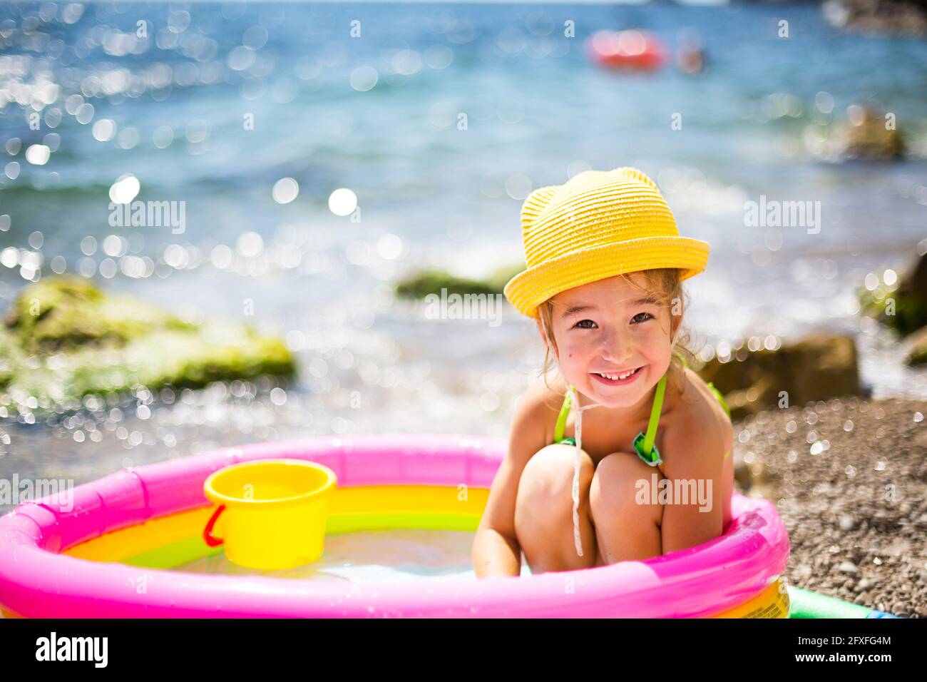 Fille en chapeau de paille jaune joue à l'extérieur près de la mer, dans  l'eau avec un seau dans une piscine gonflable sur la plage. Produits  indélébiles pour protéger les enfants Photo