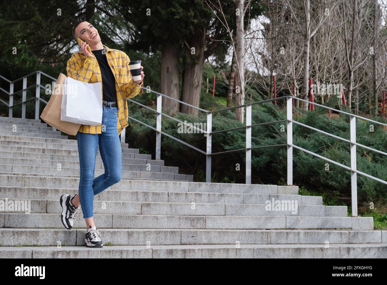 Jeune personne non binaire riant et parlant sur le smartphone en descendant les escaliers avec des sacs de shopping et une tasse de café. Faites le maquillage de la personne androgyne. Banque D'Images