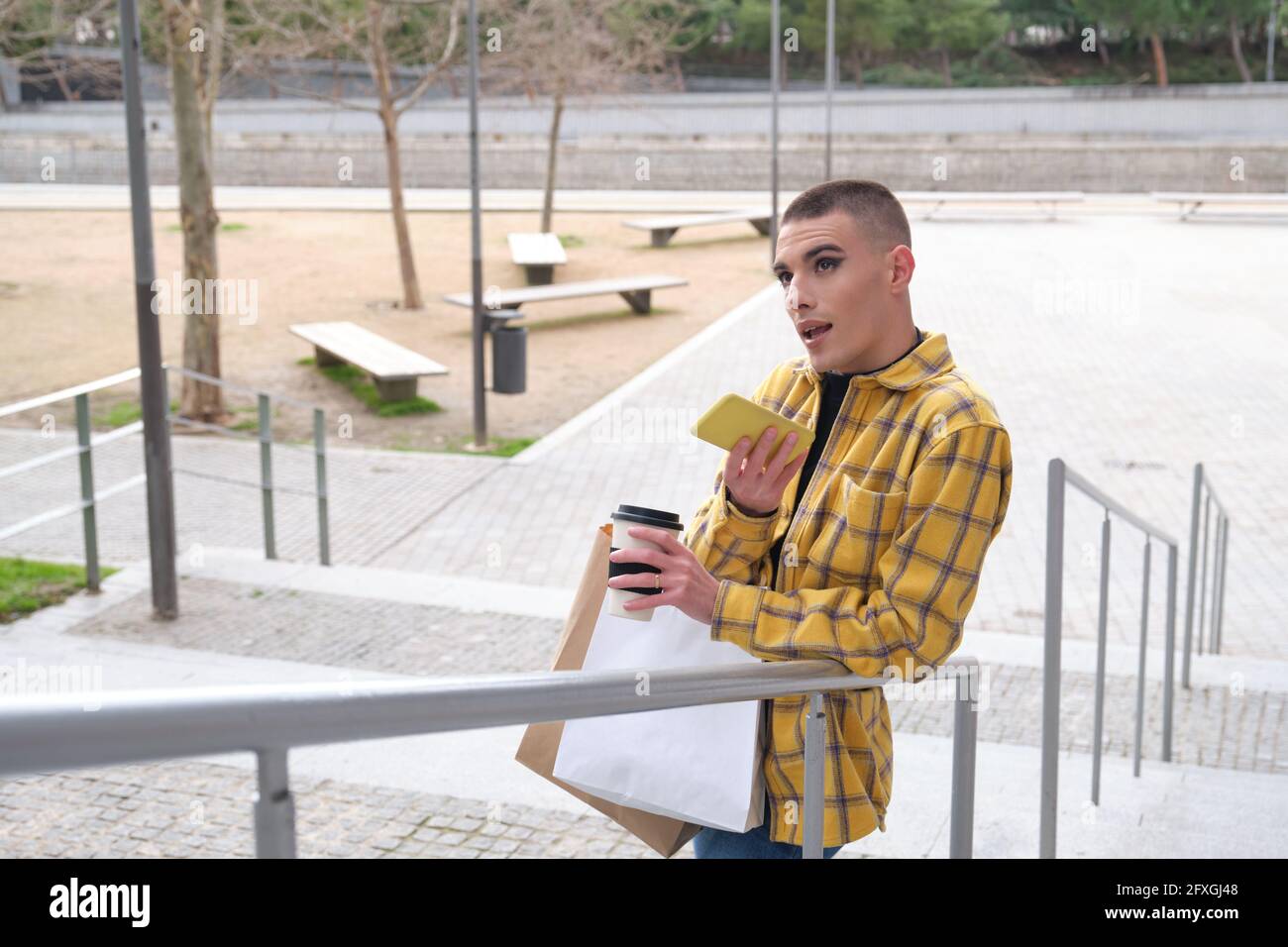 Jeune personne non binaire enregistrant un son sur le smartphone avec des sacs d'achats et une tasse de café. Faites le maquillage de la personne androgyne. Banque D'Images