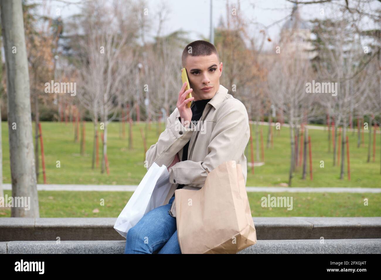 Jeune homme portant un maquillage tenant des sacs de shopping et parlant sur un smartphone assis dans un parc. Queer personne. Banque D'Images