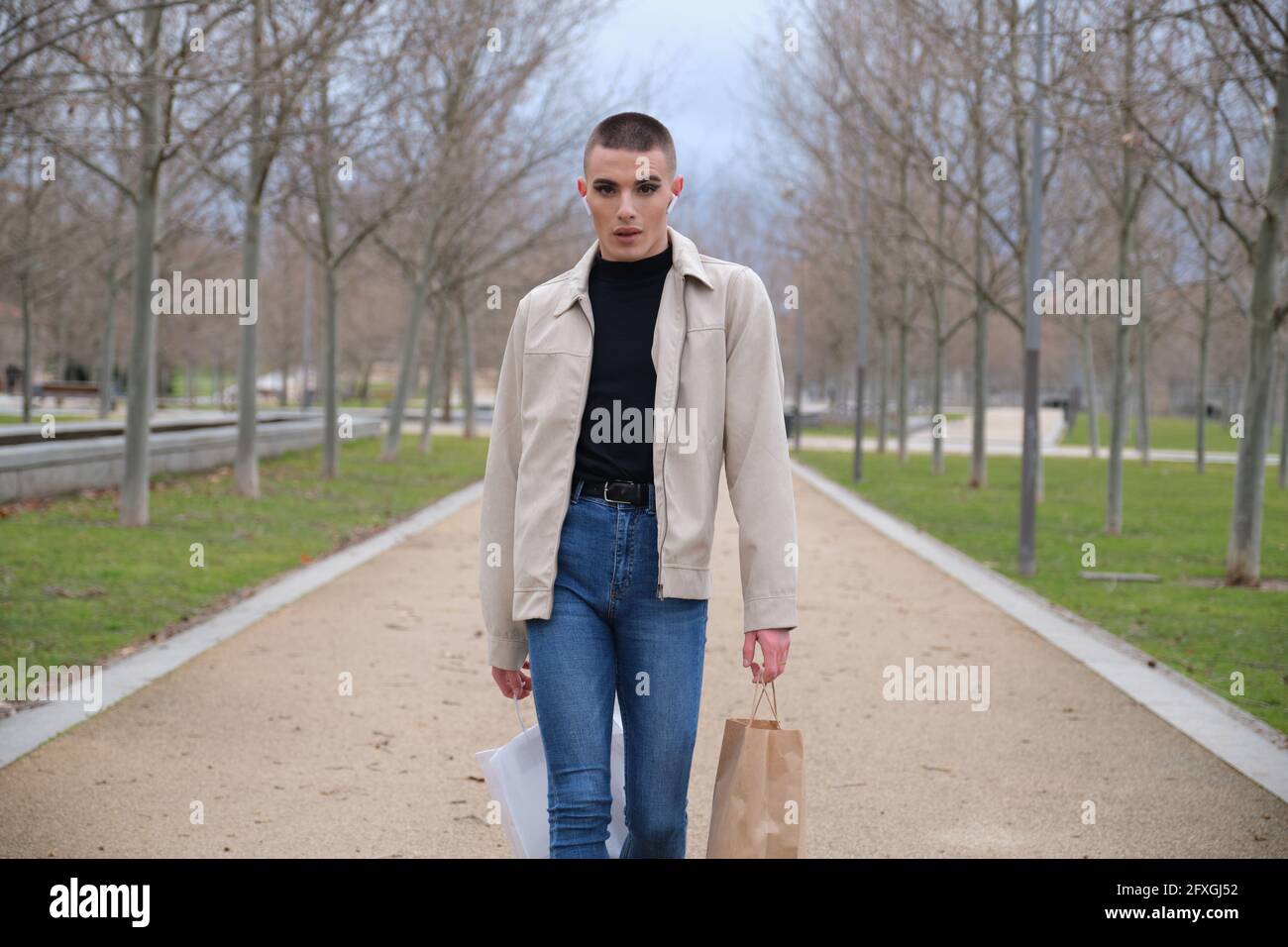 Un jeune homme portant du maquillage marche avec des sacs de shopping et écouter de la musique dans un parc. Queer personne. Banque D'Images